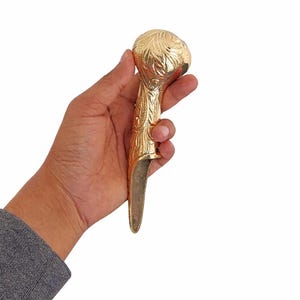 以下が含まれることがあります： 金色の金属製のオブジェクトで、装飾的な花の模様が施されています。 オブジェクトは丸みを帯びた上部と長く細いベースを持っています。 オブジェクトは白い背景に対して手で持たれています。