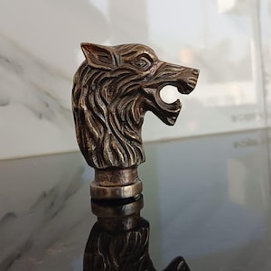 May include: A bronze bottle stopper with a detailed wolf head design. The wolf's head is sculpted with intricate fur details and an open mouth. The stopper has a metallic finish and is designed for wine bottles.