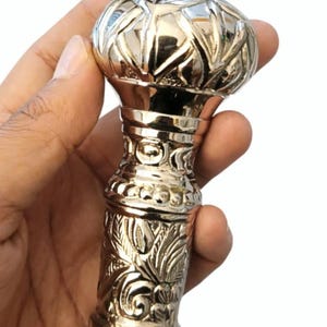 May include: A silver-coloured walking cane handle with an ornate design. The top is rounded with a textured pattern, and the shaft features floral and geometric engravings. The handle is held in a hand against a white background.