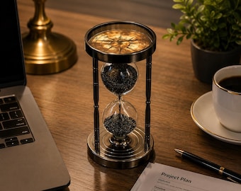 Op de afbeelding: Een antieke messing zandloper met een kompasontwerp. De glazen bollen bevatten donker zand. De zandloper staat op een houten bureau naast een laptop, een kopje koffie en een pen. De zandloper is een decoratief item, ongeveer 15 cm hoog.