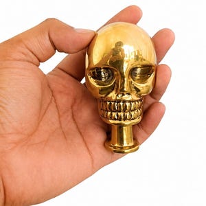 Puede incluir: Un objeto de calavera de metal dorado pulido con una superficie lisa y reflectante. La calavera presenta cuencas oculares detalladas, una cavidad nasal y una hilera de dientes definida, unida a una base cilíndrica en la parte inferior.