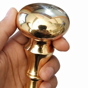 Może przedstawiać: Błyszczący, złoty uchwyt do szafki z zaokrąglonym wierzchem i dekoracyjną podstawą. Uchwyt jest trzymany w dłoni na białym tle, podkreślając jego wypolerowaną powierzchnię i design.