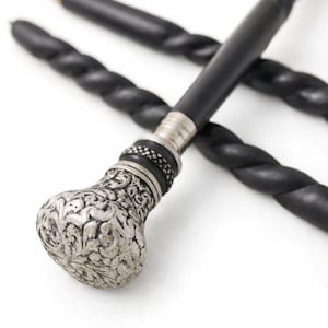 May include: Close-up of a decorative walking cane with a silver-colored, ornate handle. The handle features a detailed floral design. The shaft is black with a twisted rope-like texture. The cane is set against a white background.