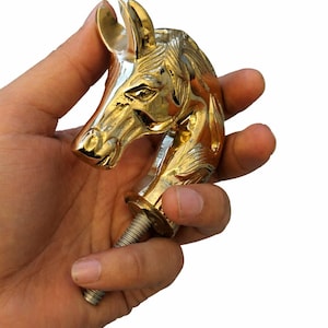May include: A golden horse head sculpture with intricate details, held in a hand. The sculpture features a threaded bolt at the base, suggesting it may be a decorative element for furniture or a similar item.