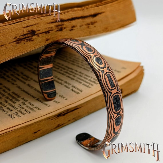 Hand-forged carbon copper Damascus bangle where layered metals are folded and twisted into a genuine ladder-wave pattern — actual blacksmithing craft you can see in every ripple.