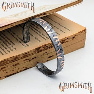 May include: Damascus steel cuff bracelet with copper accents, resting on an open book. The bracelet features a swirling pattern and copper-colored inlays. The word "GRIMSMITH" is displayed in a stylized font.