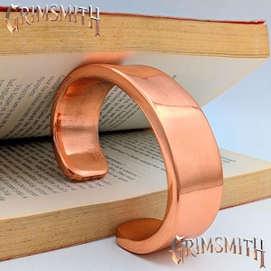 May include: A wide, polished copper cuff bracelet with a smooth, reflective surface and an open-ended design. The bracelet is positioned against an open book with visible printed text, and the word GRIMSMITH is displayed in the image.