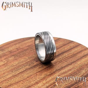 Pode incluir: Um anel prateado com uma banda estampada e uma borda lisa e polida. O anel é exibido em uma superfície de madeira. A palavra "GRIMSMITH" é visível na imagem.