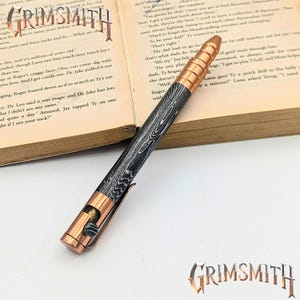 May include: A handcrafted pen with a woodgrain pattern and copper accents. The pen features a copper clip and tip, and the brand name "GRIMSMITH" is visible. The pen is resting on an open book.