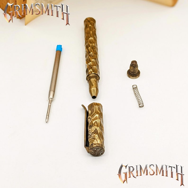 Peut inclure: Stylo en laiton d&eacute;mont&eacute; avec un corps, un clip et une pointe textur&eacute;s. Une cartouche d'encre &agrave; pointe bleue, un ressort et un bouchon &agrave; vis sont &eacute;galement visibles. Le stylo est marqu&eacute; du mot "GRIMSMITH" dans une police stylis&eacute;e.