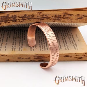 May include: A copper-colored cuff bracelet with a textured surface, resting on an open book. The bracelet has a simple, open design. The word "GRIMSMITH" is displayed above and below the bracelet.
