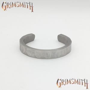 May include: A silver-colored cuff bracelet with a textured surface. The bracelet has a hammered appearance and is open at the ends. The word "GRIMSMITH" is displayed in a stylized font above and below the bracelet.