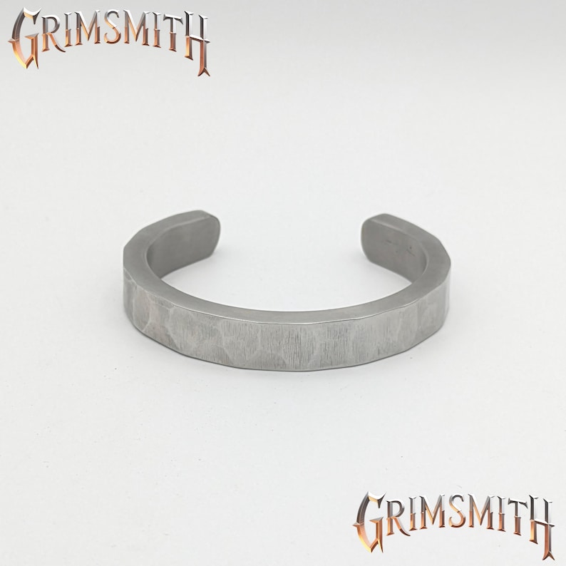 May include: A silver-colored cuff bracelet with a textured surface. The bracelet has a hammered appearance and is open at the ends. The word "GRIMSMITH" is displayed in a stylized font above and below the bracelet.
