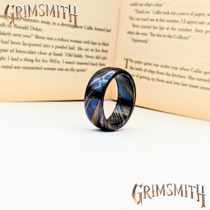 May include: A ring with a unique design, featuring a blend of blue, gold, and black colors. The ring is set against an open book. The word "GRIMSMITH" is displayed at the top and bottom of the image.