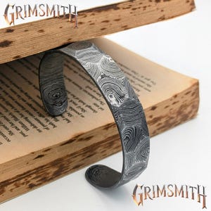 Könnte beinhalten: Ein strukturiertes Metallarmband mit einem einzigartigen Muster, das auf einem offenen Buch liegt. Das Armband hat ein gebogenes Design und besteht aus einem dunklen, gemusterten Metall. Das Wort "GRIMSMITH" wird in einer stilisierten Schriftart angezeigt.