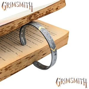 May include: A patterned steel cuff bracelet with a gold-coloured chevron design. The bracelet is displayed on an open book. The Grimsmith logo is visible in the top left and bottom right corners.
