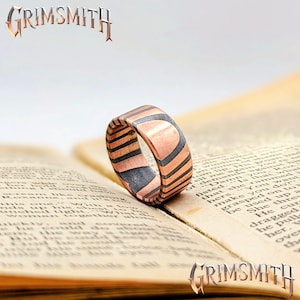 May include: A copper-toned ring with a geometric design of alternating black and copper stripes. The ring is set against the backdrop of an open book. The word "GRIMSMITH" is displayed in a stylized font.