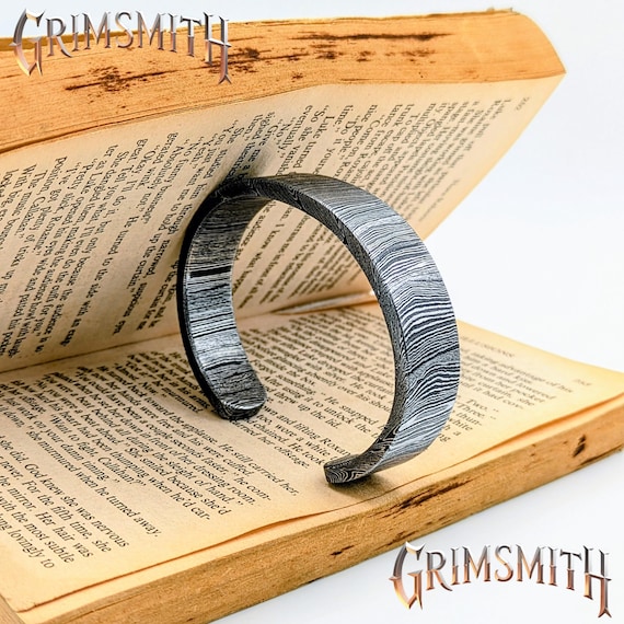 A hand-forged Damascus steel cuff bangle — the layered, flowing pattern of real Damascus metalwork makes every single piece visually one-of-a-kind in a way no factory can replicate.