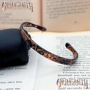May include: A cuff bracelet featuring a dark, textured metal surface with intricate, swirling copper-coloured patterns. The open-ended band has a slim, curved profile and a polished finish, displaying a distinct contrast between the black and copper tones.