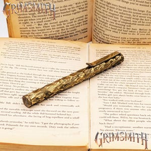 Peut inclure: Un stylo dor&eacute; avec un motif textur&eacute; unique repose sur les pages ouvertes d'un livre. Le design du stylo est complexe et les pages du livre affichent du texte imprim&eacute;. Le stylo est un instrument d'&eacute;criture.