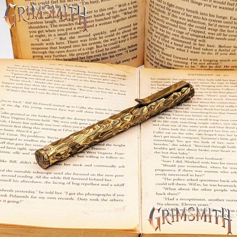 Peut inclure: Un stylo dor&eacute; avec un motif textur&eacute; unique repose sur les pages ouvertes d'un livre. Le design du stylo est complexe et les pages du livre affichent du texte imprim&eacute;. Le stylo est un instrument d'&eacute;criture.