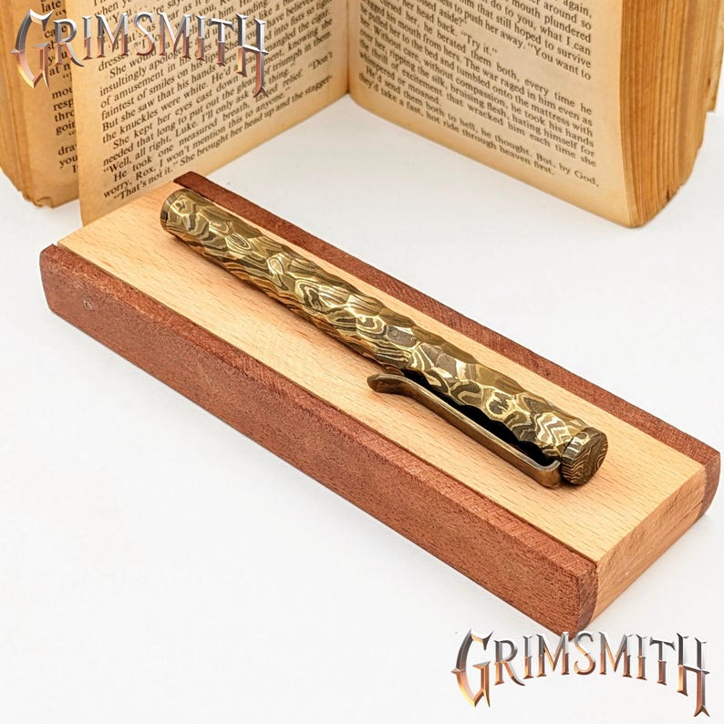 Peut inclure: Gros plan d'un stylo en laiton avec un motif textur&eacute;, pos&eacute; sur une base en bois. Le stylo a un clip et un design d&eacute;taill&eacute;. La base est faite de deux types de bois. Le logo Grimsmith est visible.