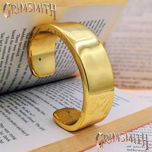 May include: A polished gold-tone metal cuff bracelet with a smooth, curved surface and an open back design. The bracelet is positioned upright, resting on the open pages of a book with the text GRIMSMITH visible at the top and bottom corners.