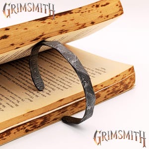 May include: A metal bookmark with a unique pattern is inserted into an open book. The bookmark has a curved design and is made of a dark, patterned metal. The book's pages are aged, and the text is visible. The brand name "GRIMSMITH" is displayed.