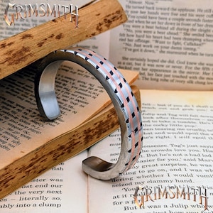 May include: A silver cuff bracelet with a geometric design featuring copper and black accents. The bracelet is displayed on an open book. The Grimsmith logo is visible in the image.