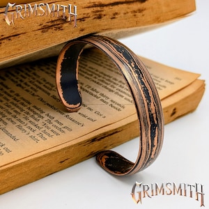 May include: A copper-toned cuff bracelet with a black wood grain pattern. The bracelet is curved and rests on the pages of an open book. The Grimsmith logo is visible in the image.