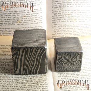 Pode incluir: Dois objetos quadrados de metal texturizado com um padrão único de grão de madeira. Os objetos são cinza escuro e estão sobre um livro vintage. A palavra "GRIMSMITH" é visível na imagem.