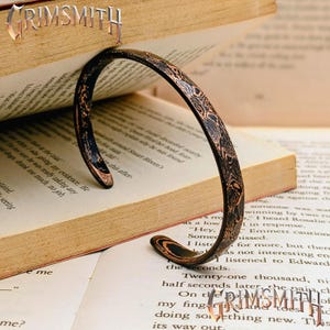 May include: A curved cuff bracelet featuring a dark, textured surface with intricate, swirling copper-coloured patterns. The metal band has an open design and displays a distinct layered aesthetic throughout its entire length and rounded edges.