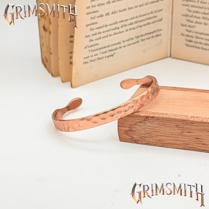 May include: A hammered copper cuff bracelet with a textured surface. The open-ended bracelet is displayed on a wooden box next to an open book. The word "GRIMSMITH" is visible in the image.