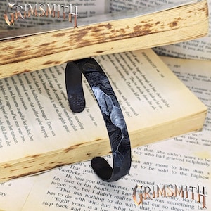 May include: Black metal cuff bracelet with a textured pattern, resting on an open book. The bracelet has a curved design and a dark finish. The word "GRIMSMITH" is visible in the upper left and lower right corners.