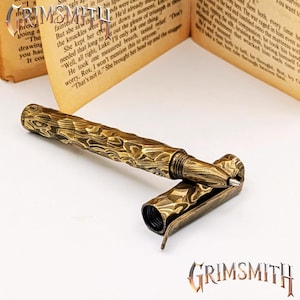 Peut inclure: Gros plan d'un stylo en laiton avec un motif textur&eacute; en forme de grain de bois. Le capuchon du stylo est ouvert, r&eacute;v&eacute;lant la pointe. Le stylo est pos&eacute; sur un fond flou d'un livre ouvert. Le logo Grimsmith est visible.