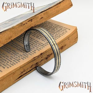 May include: A Damascus steel cuff bracelet with two gold-colored stripes is displayed open on a vintage book. The bracelet has a textured, patterned surface. The word "GRIMSMITH" is displayed in a stylized font.