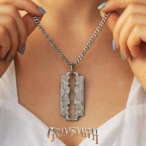 Pode incluir: Um colar prateado com um pendente em forma de lâmina de barbear. O pendente da lâmina tem um design padronizado único e pende de uma corrente. A palavra "GRIMSMITH" é exibida em uma fonte estilizada na parte inferior da imagem.