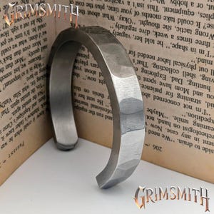 May include: A silver-colored, hammered metal cuff bracelet. The bracelet has a faceted design and a curved, open shape. The word "GRIMSMITH" is visible in the image.