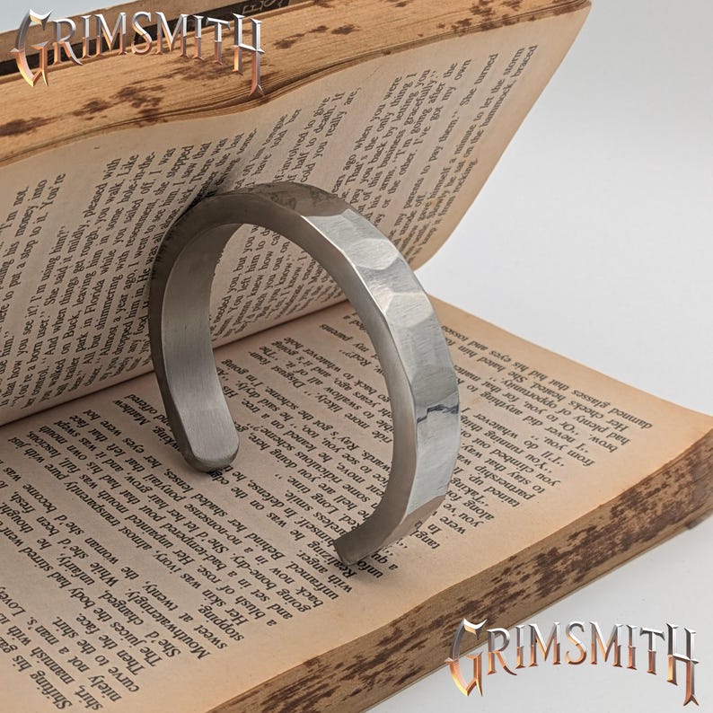 May include: A silver-colored, hammered metal cuff bracelet. The bracelet has a geometric, faceted design and is displayed open on the pages of an old book. The word "GRIMSMITH" is visible in the image.