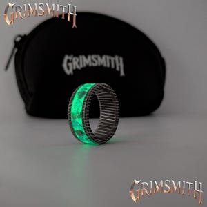 May include: A glowing green ring with a dark metal band. The ring has a unique design with a bright green inner band. The word "GRIMSMITH" is displayed in a gold font at the top and bottom of the image, and on a black pouch in the background.