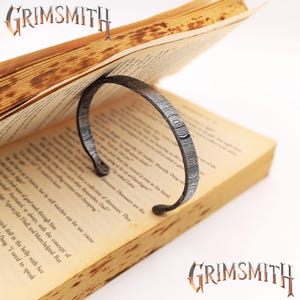 May include: A patterned metal book page holder supporting an open book. The book's pages are aged with printed text. The Grimsmith logo is displayed in the top left and bottom right corners.