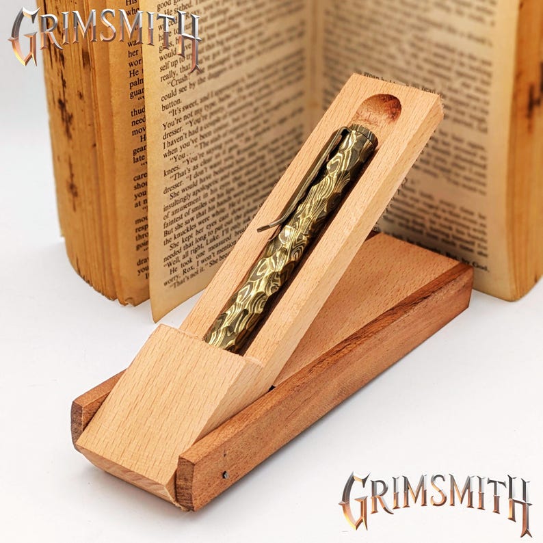 Peut inclure: Un stylo en laiton dor&eacute; avec un motif est pr&eacute;sent&eacute; dans un coffret en bois ouvert. Le stylo a une surface textur&eacute;e. Le coffret est en bois clair. Un livre ouvert est en arri&egrave;re-plan.