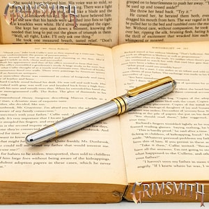 May include: A silver and gold-toned fountain pen with a ribbed design, resting on an open book. The pen has a gold clip and accents, and the book's pages are yellowed with text. The pen is a writing instrument.