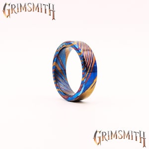 May include: A colorful ring with a unique pattern of blue, gold, and brown swirls. The ring is made of metal and has a smooth, polished finish. The word "Grimsmith" is displayed above and below the ring.