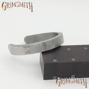 May include: A silver-colored cuff bracelet with a textured surface rests on a dark brown block. The bracelet has a hammered appearance and an open design. The word "GRIMSMITH" is displayed in a stylized font above and below the bracelet.