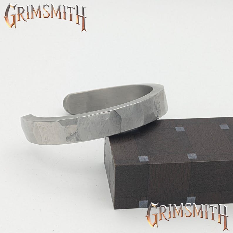 May include: A silver-colored cuff bracelet with a textured surface rests on a dark brown block. The bracelet has a hammered appearance and an open design. The word "GRIMSMITH" is displayed in a stylized font above and below the bracelet.