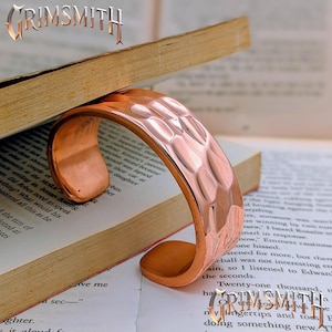 May include: A wide, open-ended copper cuff bracelet featuring a hammered, textured surface with reflective highlights. The metal has a warm, reddish-brown metallic finish. The word GRIMSMITH is visible in the top left and bottom right corners.