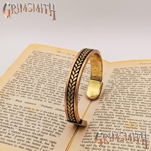 May include: A copper and brass cuff bracelet with a wheat pattern design. The bracelet is open, resting on a vintage book with text. The Grimsmith logo is visible.