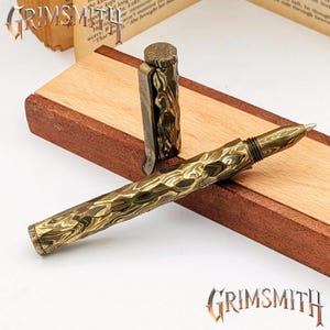 Peut inclure: Gros plan d'un stylo au motif textur&eacute; unique, dans des tons or et marron. Le capuchon du stylo est ouvert, r&eacute;v&eacute;lant la pointe. Le stylo repose sur une surface en bois avec une bordure marron fonc&eacute;. Le logo Grimsmith est visible.
