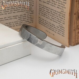 May include: A silver-toned cuff bracelet with a hammered texture. The open-ended design features a flat, wide band with a slightly curved shape. The bracelet is displayed on a white surface with a book in the background. The Grimsmith logo is visible.